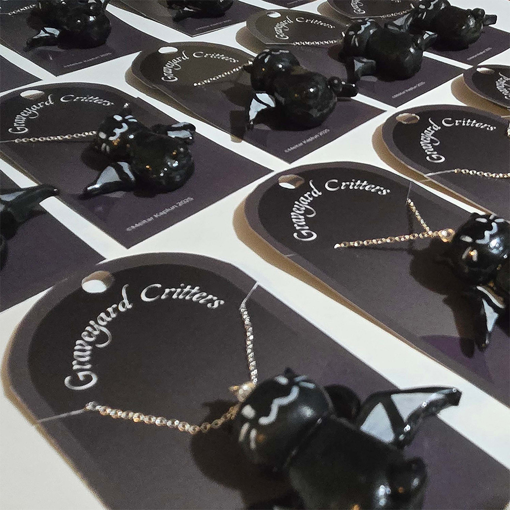 Close-up view of multiple black cat-bat necklaces displayed on individual black tags labeled 
                        Graveyard Critters, arranged neatly in rows.