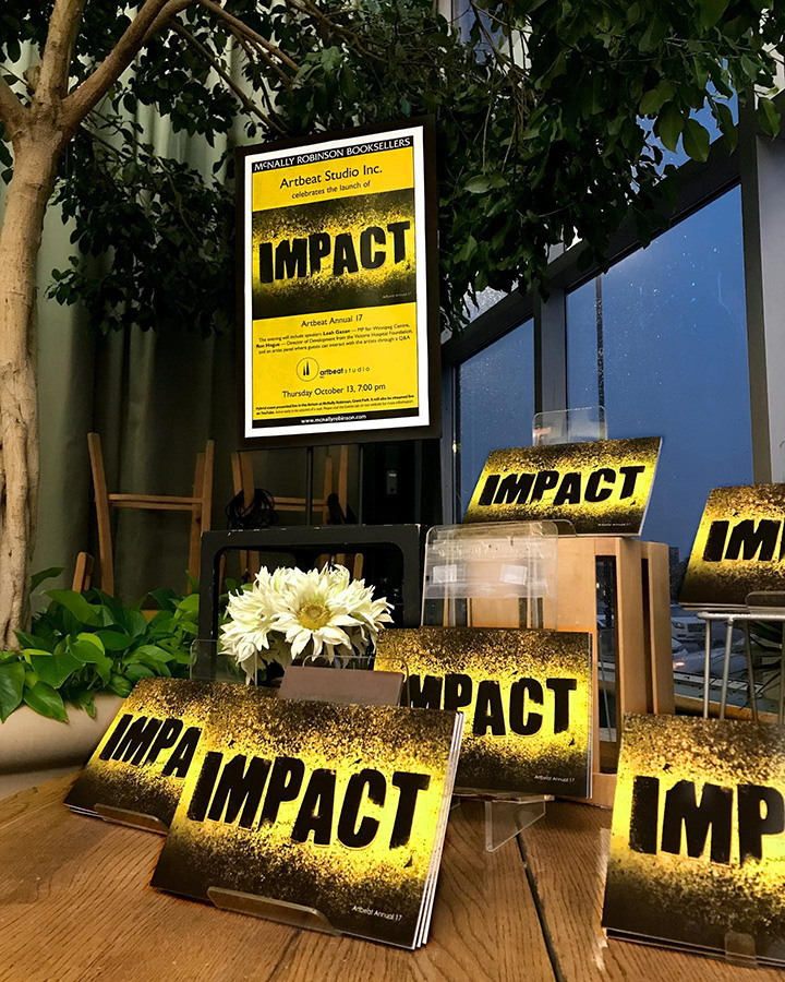 This image shows an indoor display for the IMPACT Artbeat Annual 17 book launch at McNally Robinson Booksellers. The display includes a poster with the word IMPACT prominently featured, along with the event details. Several copies of the books and white flowers are arranged around the poster. In the background is an artificial tree and a window.