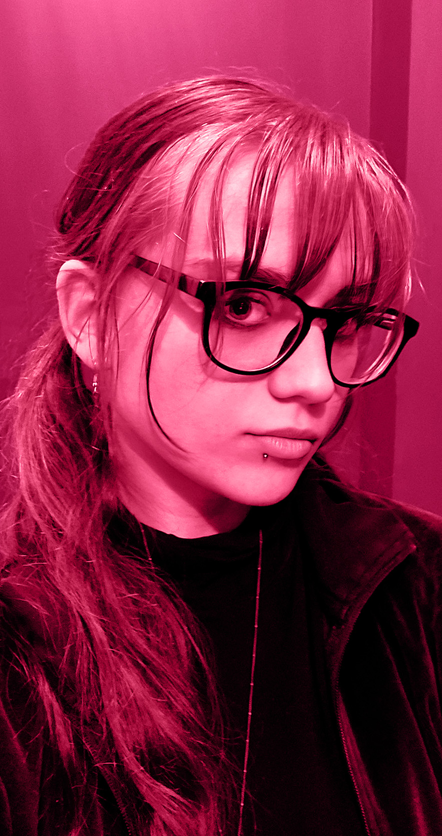 A close-up portrait shows a person with light skin, long medium coloured hair with low pigtails, black-framed glasses, and two lip piercings. The person is wearing a dark top, and the image has a strong pink hue.